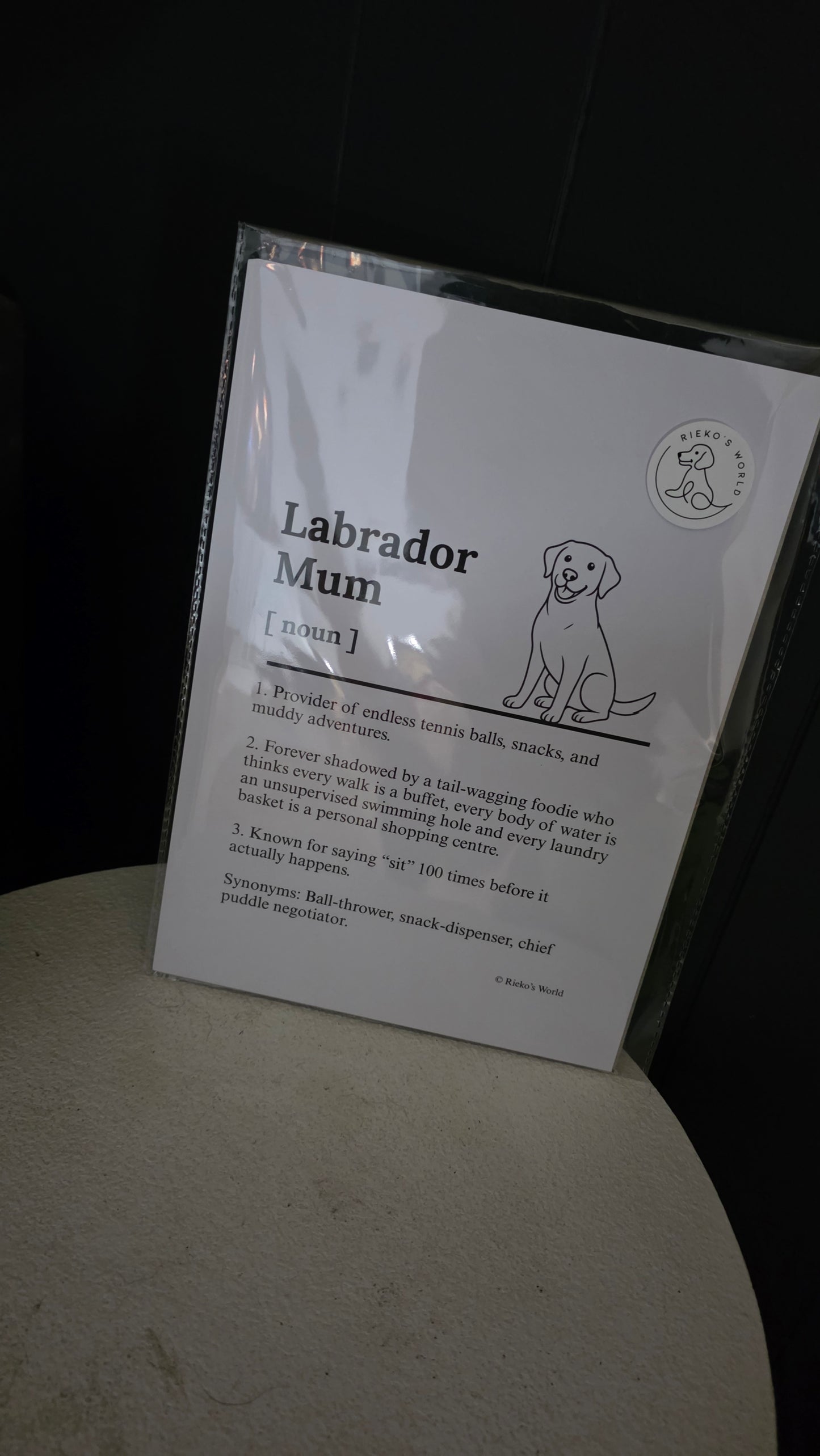 A printed card titled "Labrador Mum" with a small dog illustration and text, sealed inside a clear plastic sleeve and leaning on a light surface against a dark background.