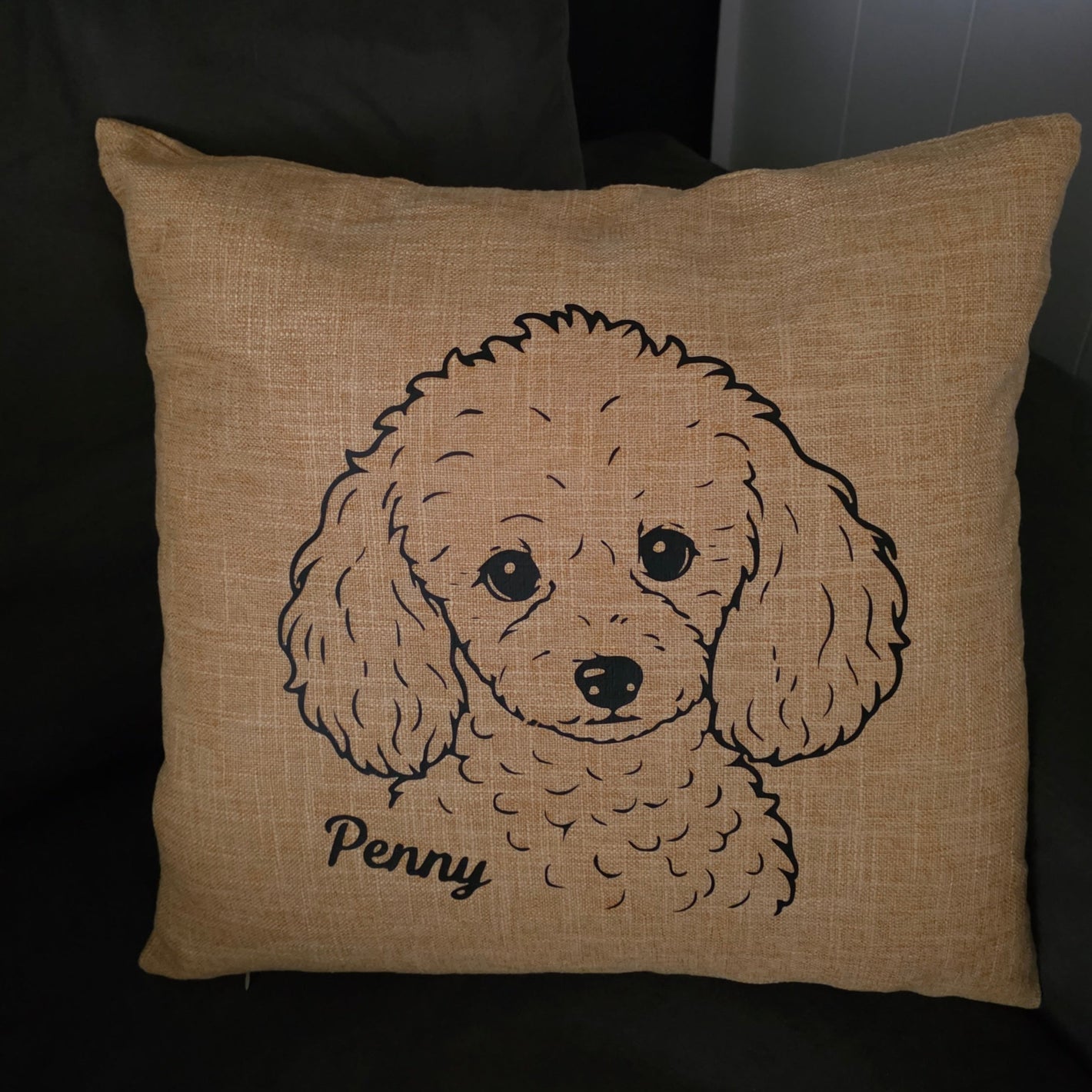 A square tan burlap-style throw pillow featuring a black line art illustration of a poodle dog and the name 'Penny' in script, resting on a dark couch.