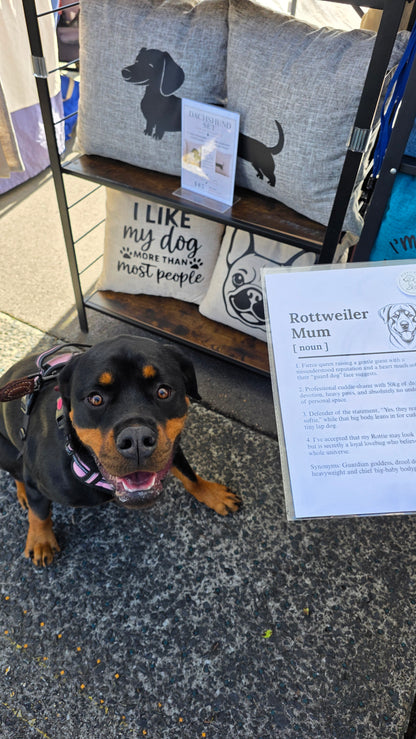 A Rottweiler dog wearing a pink harness sits on a gray pavement next to a display stand featuring dog-themed pillows and a sign labeled 'Rottweiler Mum [moon]'.