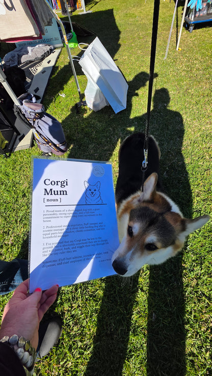A person holding a printed 'Corgi Mum [200m]' paper while a corgi dog on a leash looks on outdoors on grass.