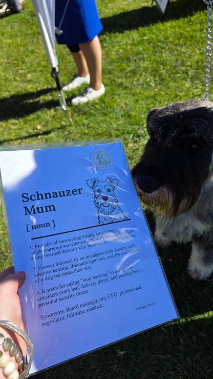 A person holding a blue printed card titled 'Schnauzer Mum' outdoors on grass, with a black-and-tan dog standing beside it. The card includes a small dog illustration and dictionary-style definition text.