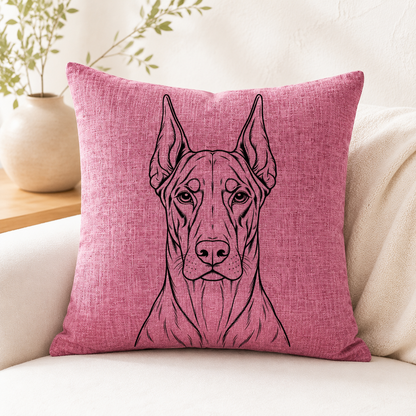 A square pink throw pillow with a black line art illustration of a Great Dane dog, resting on a cream-colored sofa with a neutral background and a potted plant.