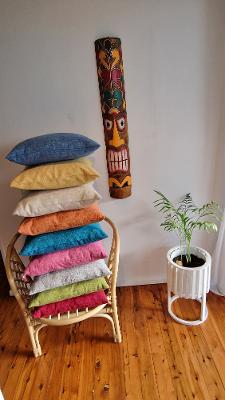 A stack of colorful throw pillows in shades of blue, yellow, cream, orange, pink, teal, green, and purple rests on a wicker chair next to a tall wooden tiki mask and a potted plant against a plain wall.