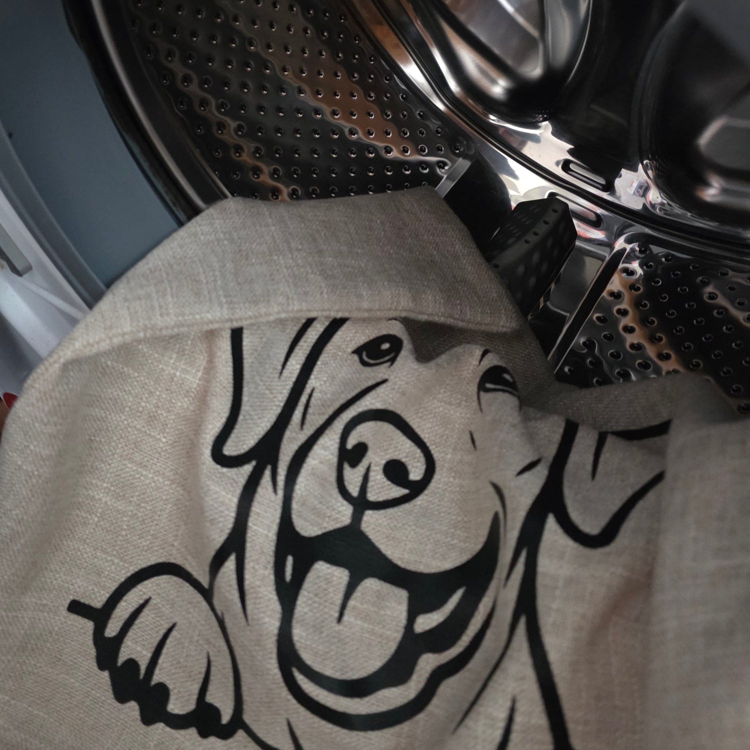 A close-up of a beige cushion cover with a black labrador outline dog graphic, partially pulled out of a front-loading washing machine drum made in Australia by Rieko's World.