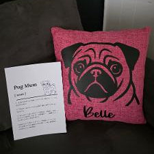 A pink throw pillow with a black pug face graphic and the name 'Belle' printed on it, displayed next to a white paper tag on a dark couch. Personalised name Pug pink cushion and breed print by Rieko's World