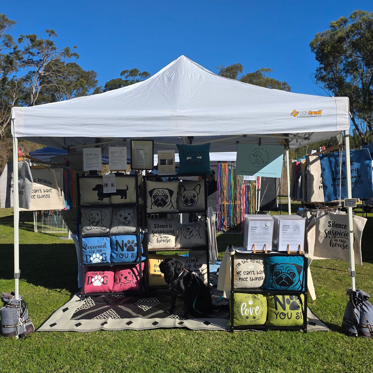 A white pop-up canopy tent set up outdoors on a grassy lawn under a clear blue sky, displaying various printed dog cushions and prints made by Rieko's World