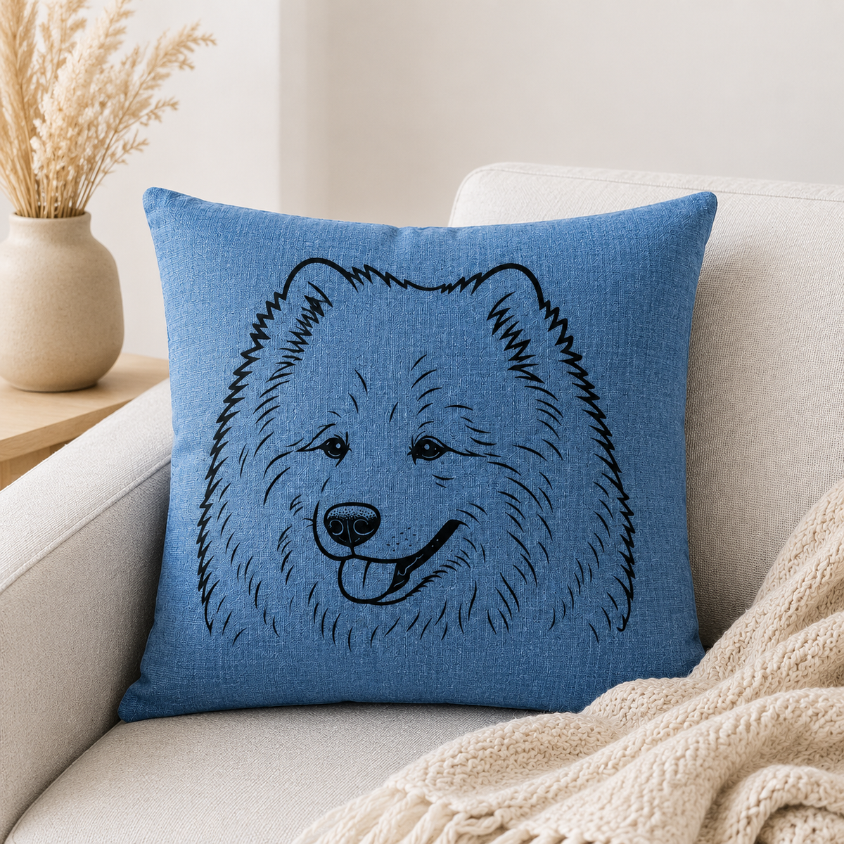 A square blue throw pillow with a black line-art illustration of a fluffy dog face, resting on a light beige sofa with a cream knit blanket and a vase of dried grass in the background.