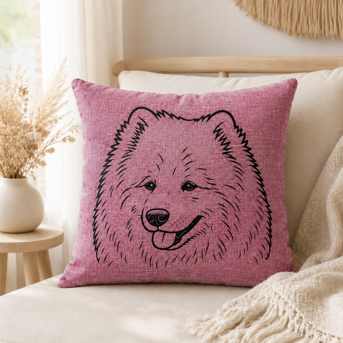 A square pink throw pillow with a black line-art illustration of a fluffy dog face, styled on a cream sofa with a beige throw blanket and dried flowers in a vase.