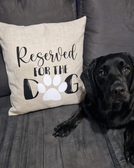 A close up of a beige cushion cover with 'Reserved for the Dog' text and a paw print design on grey sofa with black labrador sitting next to it made in Australia by Rieko's World.